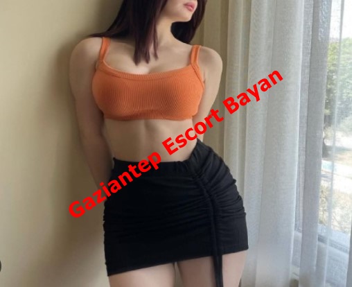 Woman in an orange cropped top and black ruched mini skirt standing by a window, with red diagonal text 'Gaziantep Escort Bayan' across the image.