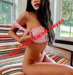 Woman with long dark hair sits on a striped couch by a window, wearing a pink sheer top (revealing outfit).